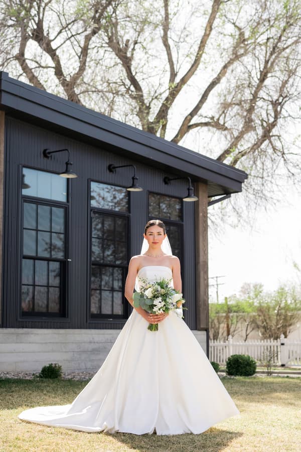 A bride is standing outside of a house holding flowers.