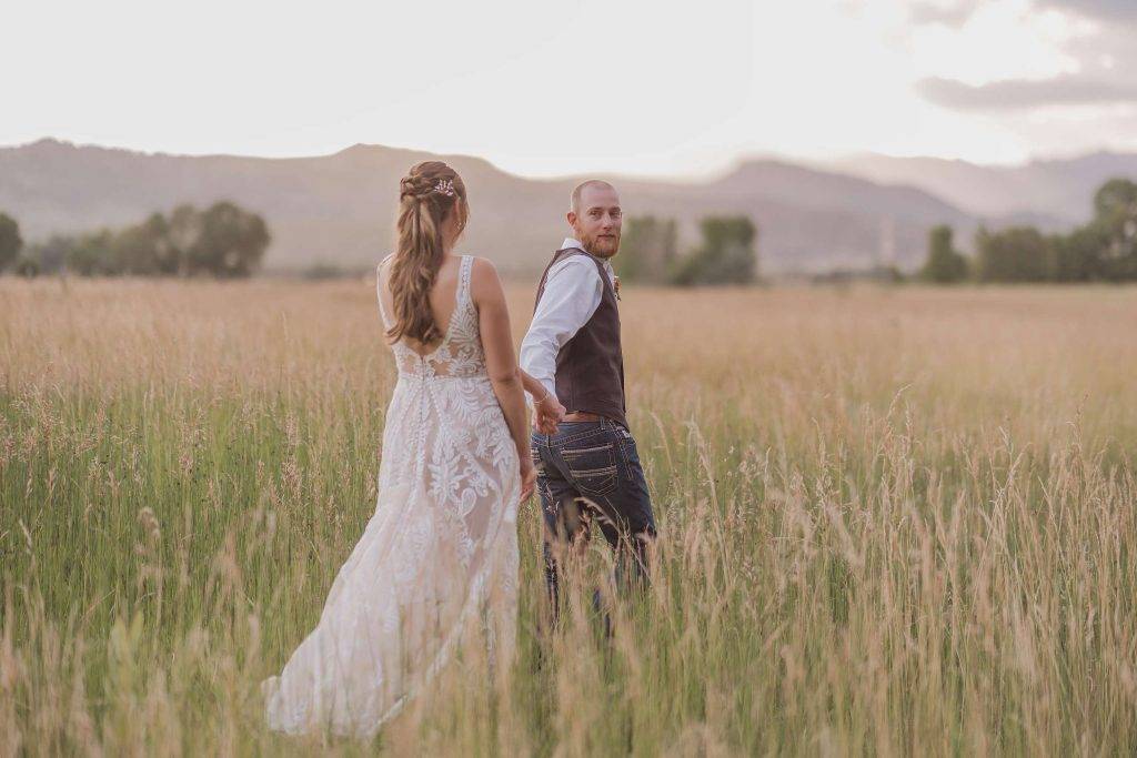 A groom guiding is bride through tall grasses in the foothills of Colorado