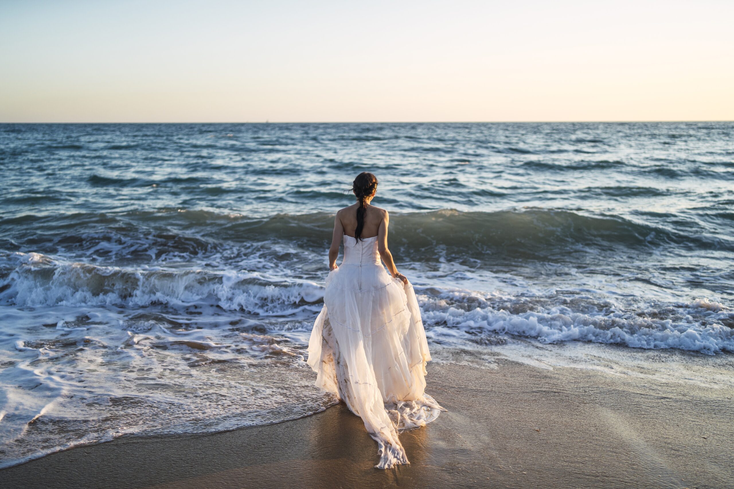 Brunette caucasian bride approaching the sea in a white wedding dress
